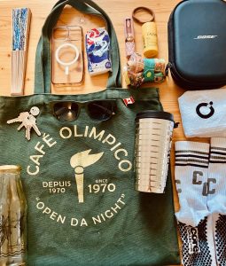 A green tote bag from a café in Montreal sits on a surface with its contents spread out around it: a folded reusable bag, a hand fan, a pack of tissues, a lip gloss, sunscreen, a phone case, a pair of headphones, a gym towel, grippy socks, a Nespresso travel cup, a glass water bottle, a set of keys with a ceramic charm marked “Sept 20,” and a small Canada flag pin.