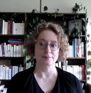 Self-portrait in office. I am in the foreground, pictured from the shoulders up. In the background are bookcases (French and medieval literature, modern literary criticism and theory, poetry and poetics). A potus plant is on top of the bookcase. Its vines entangle the shelves and sides below, and seem to grow out of my ear. Originally arranged as a background while teaching online, then photographed for use as a Zoom background. Composition and photo: Juliet O’Brien, 2024. 