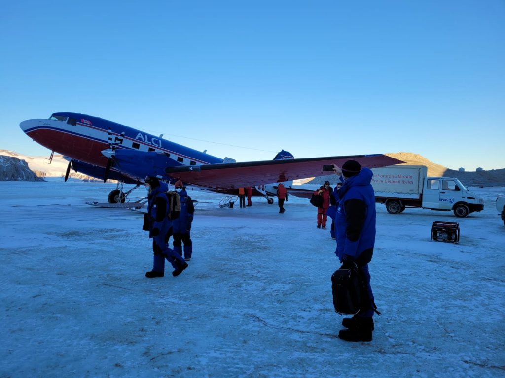 Image of a number of scientists and staff wearing blue and red at the station near an aeroplane which is blue and red. The sky is blue with some clouds, the weather looks quite calm. The people and plane are standing on snow covered ice. There is also a truck which is collecting materials off of the aeroplane. Mountains covered in snow can be seen in the background.