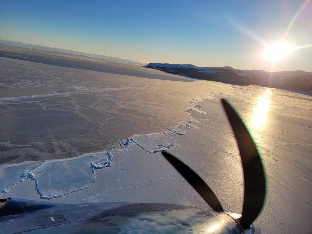 Photo taken from a plane as it flies over a large ice sheet. Mountains can be seen in the background with the plane propellor in the front.