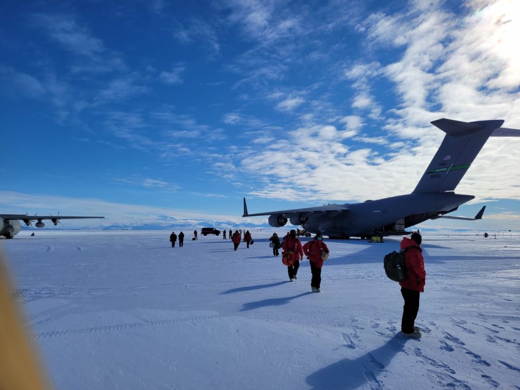 Image of a number of scientists and staff wearing red at the station near an aeroplane. The sky is blue with some clouds, the weather looks quite calm. The people and plane are standing on snow covered ice.
