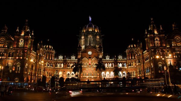 Busy night time photograph of front view of Gothic railway station with central garden.
