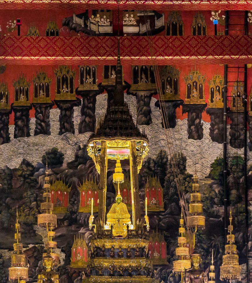 The Emerald Buddha sitting in small golden temple with nine tiered umbrella and backdrop of murals.