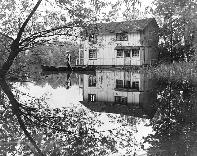 Black and white photo, reflection of boathouse and boater.