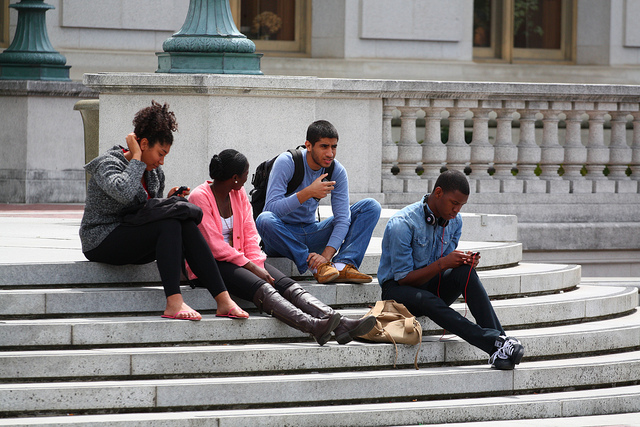 college students sitting on stone steps of a building, three of them with phones in their hands