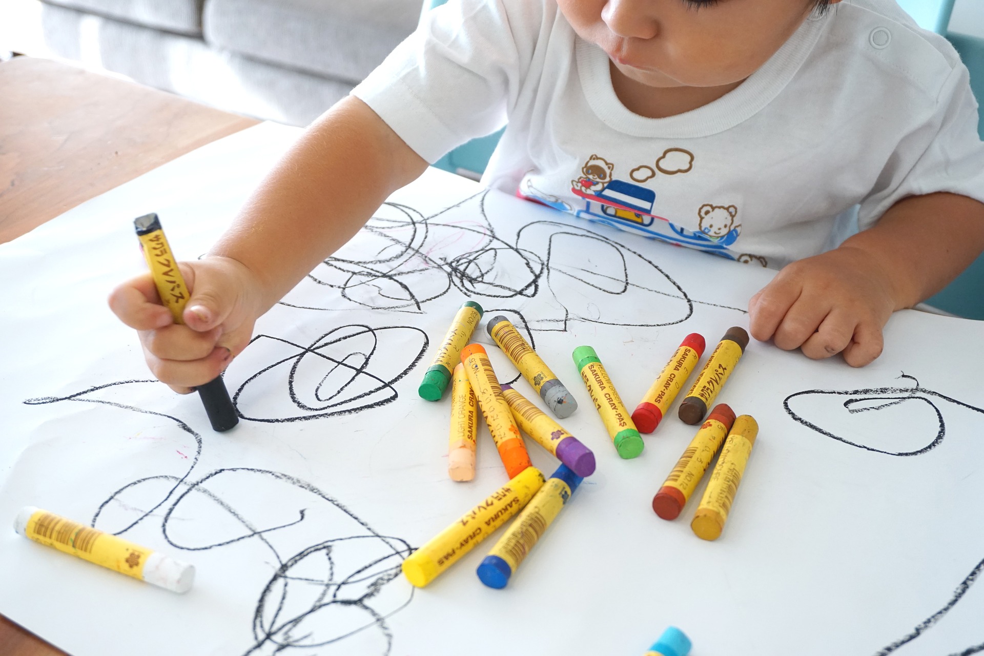 A view from above of a child scribbling with colorful crayons on a large piece of paper.