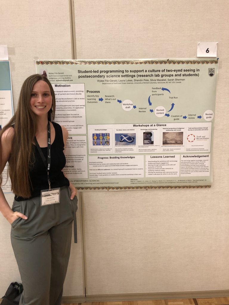 A young woman in a black sleeveless top and green trousers poses in front of the a green poster titled, ”Student-led programming to suportaculture of two-eyed seeing in postsecondary science settings (research lab group and students).”