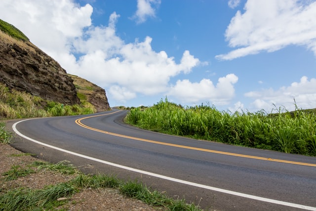 A road leading to the crest of a hill, with a mountain on one side, and tall grass on the other. The sky is bright blue with white, fluffy clouds.