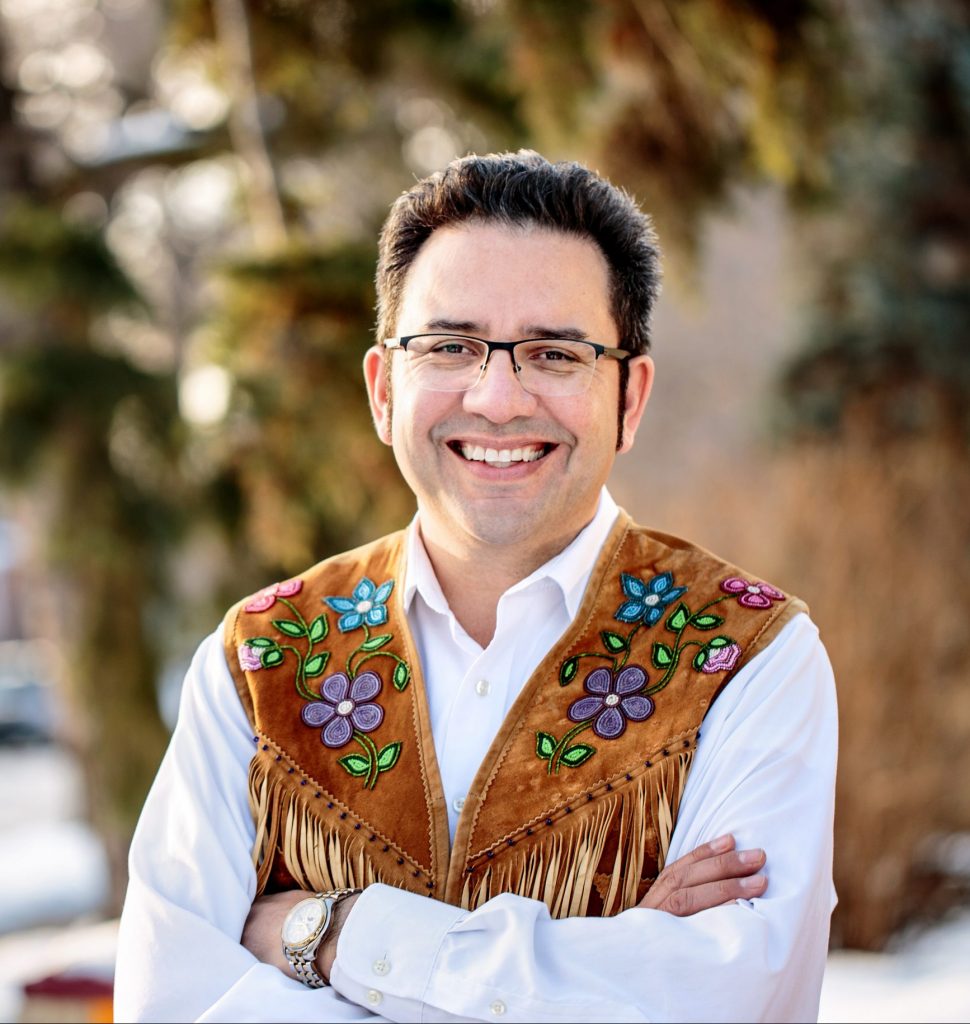 Richard Van Camp, a man in glasses and a colourfully embroidered vest stands against the backdrop of a snowy forest.