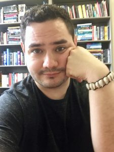 Dr. Angel Matos, a man with dark hair and eyes, rests one cheek on a fist in front of a bookshelf