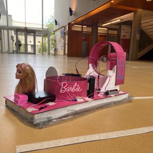 A handmade hovercraft is decorated with a Barbie theme, the body is pink with the Barbie logo and a Barbie doll head at the front as a mascot. The hovercraft has a large turbine at the back for propulsion.