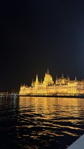 The Budapest parliament building lights up the night, seen from a boat riding down the Danube canal. The waters are calm and peaceful.