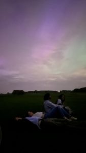 A group of people sit and lie under the sky, which is filled with views of the green and purple Northern Lights. The field they lie in is empty, and the trees lining the horizon can be barely made out in the darkness.