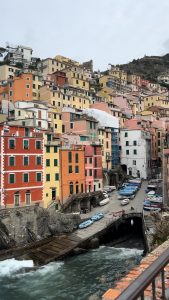 Colourful buildings rise in rows up a hillside in the town of Cinque Terre, Italy. Ocean waves crash against a dock at the bottom of a road lined with small boats.