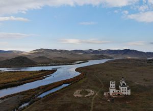 Photo of broad valley with river flowing right through it, perhaps confluence of two rivers or arms of rivers. On one of the sides sits an abandoned orthodox looking building.
