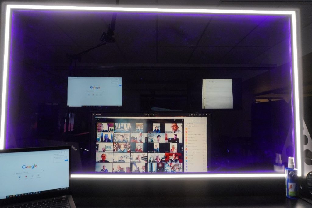 View from the professor's side of the lightboard. Three monitors are seen through the lightboard, with one monitor displaying a Zoom class.
